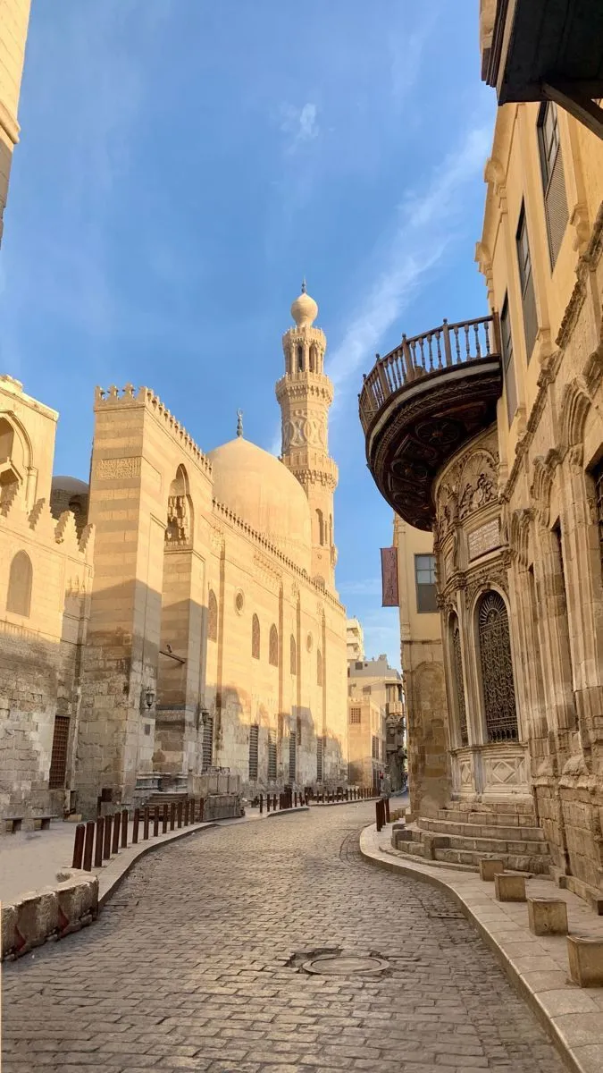 شارع المعز في القاهرة الإسلامية وقت الغروب، وطريقه المرصوف يمتد بين جدران المساجد التاريخية ومئذنة ومشربية خشبية