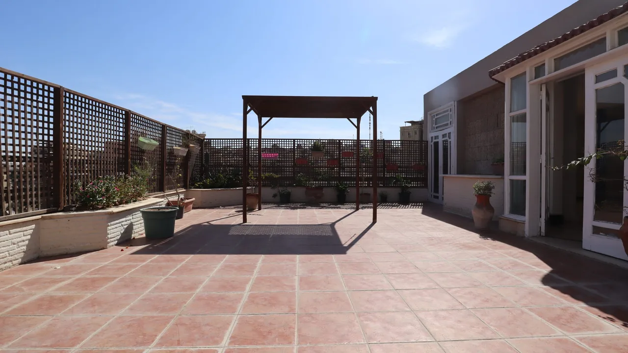 Wide tiled roof terrace with a metal pergola frame, screened walls and planted borders, at a penthouse for rent in Degla
