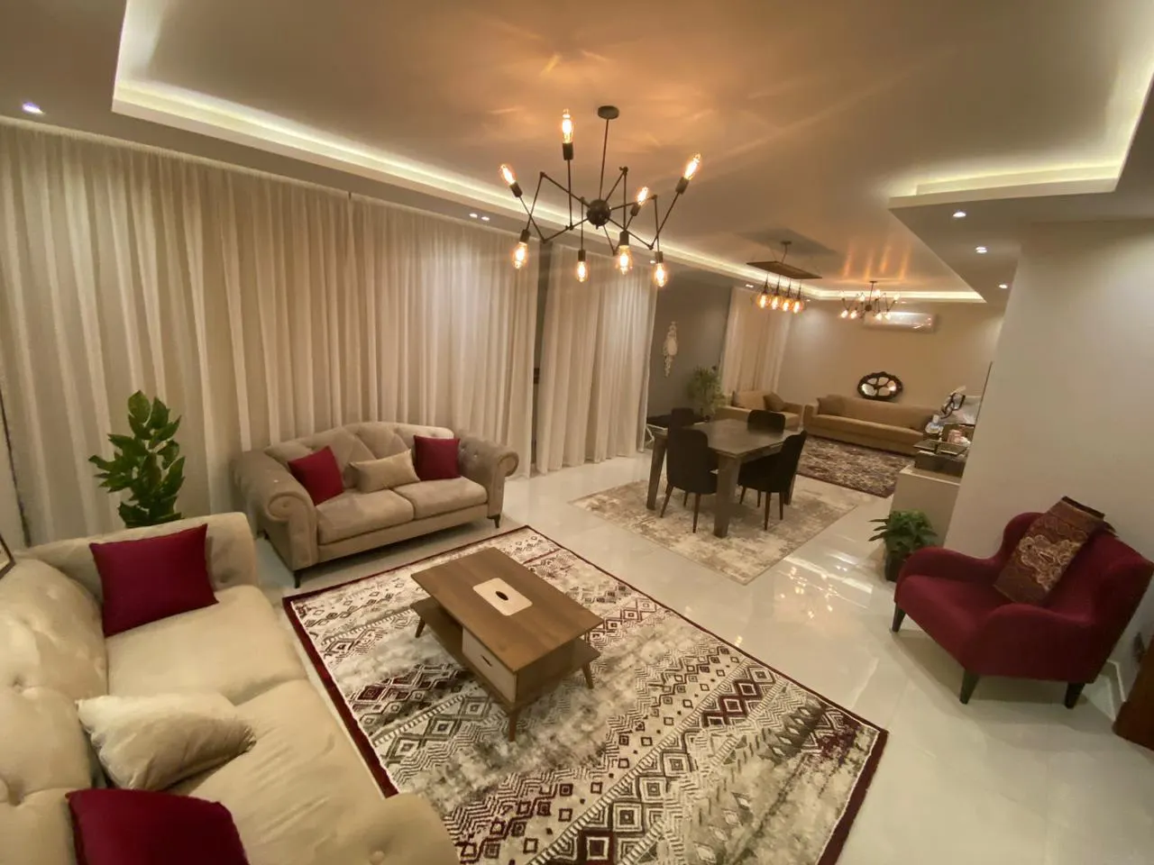 Spacious reception of a 360 m² apartment in New Cairo with beige sofas, red armchairs and a dining area behind