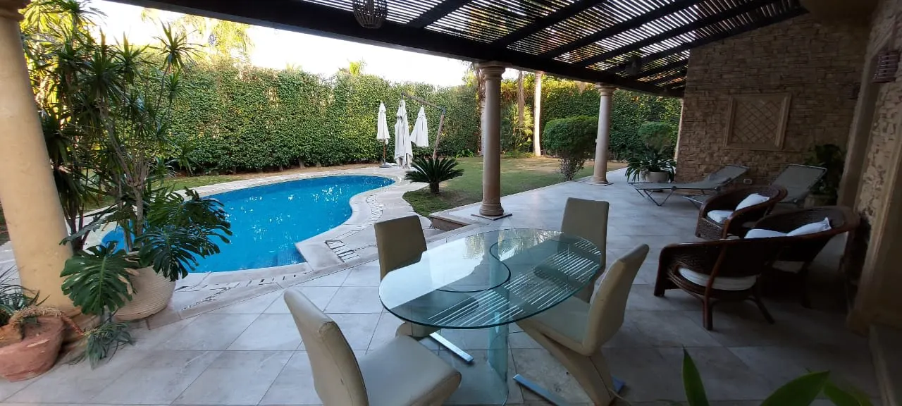 Pergola-shaded terrace with a glass dining table beside the pool and private garden of an apartment in Katameya Heights