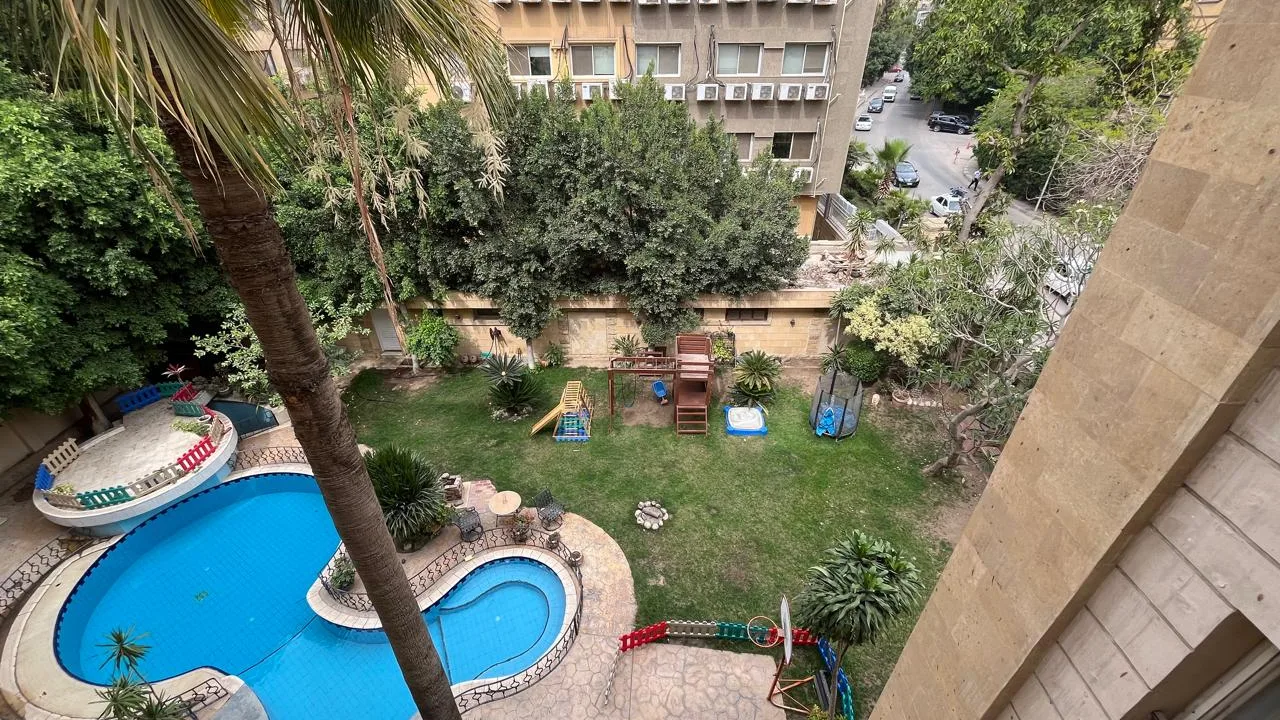 حديقة مشتركة بحمام سباحة منحني ونجيل ونخيل وألعاب أطفال، مرئية من دور علوي في شقة للإيجار في سرايات المعادي