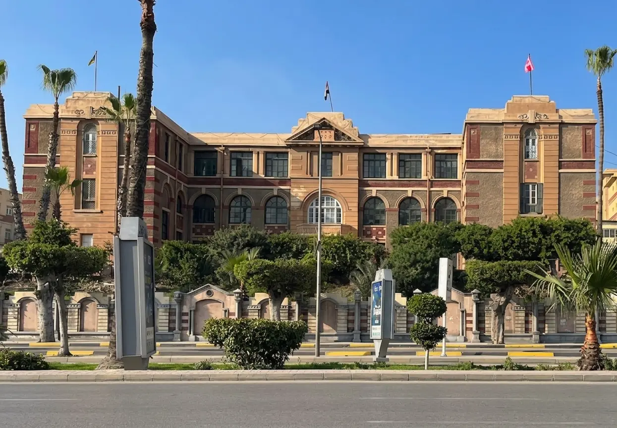 الواجهة الحجرية الطويلة المخططة لمدرسة النصر للبنين في الزمالك خلف أشجار النخيل وسور الحديقة، ويرفرف فوقها العلم المصري، القاهرة