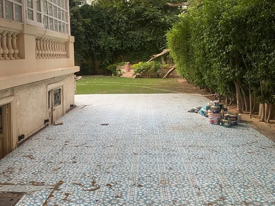 فناء مبلط وحديقة بجوار مبنى كلاسيكي على طراز الفيلات تحيط بهما أسوار نباتية وأشجار عالية في دور أرضي في الزمالك